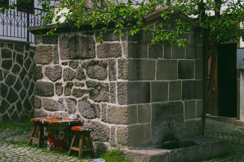 A rustic outdoor stone fountain with wooden stools on a cobblestone path, surrounded by greenery.
